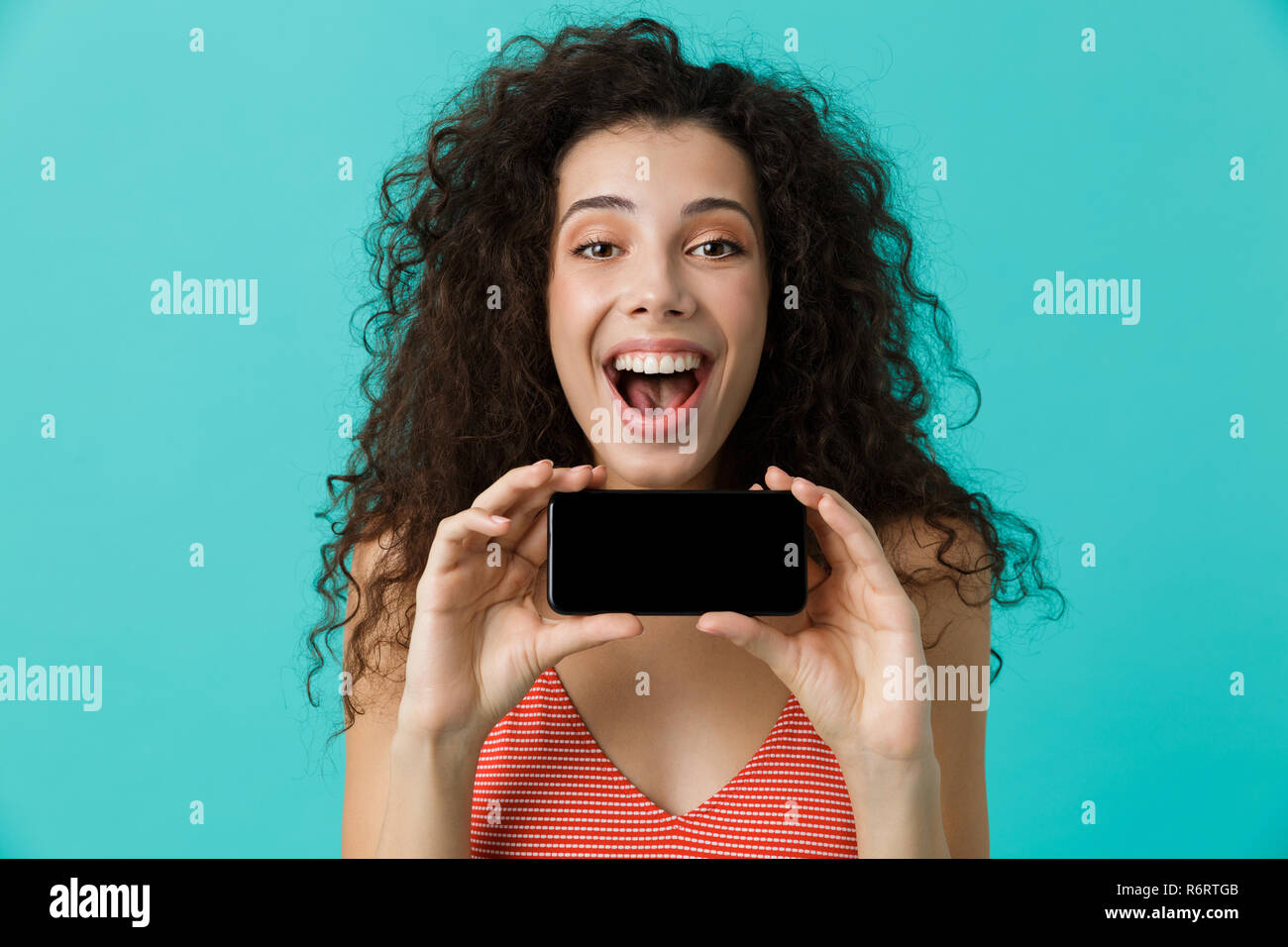 Photo de femme excité 20s avec des cheveux bouclés smiling and holding mobile phone isolé sur fond bleu Banque D'Images