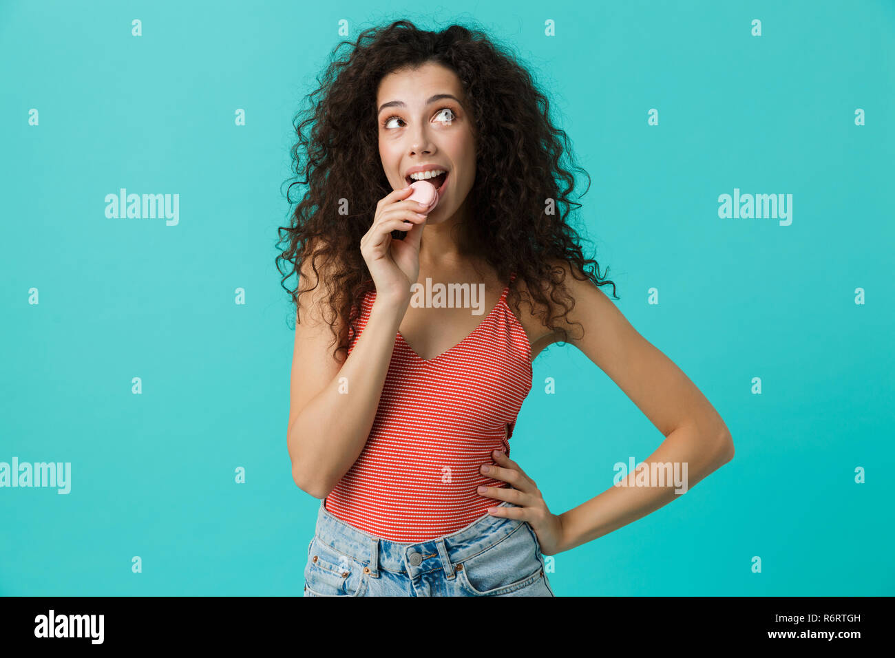 Photo de jolie femme 20s avec les cheveux bouclés de sourire et de manger biscuit macaron isolé sur fond bleu Banque D'Images