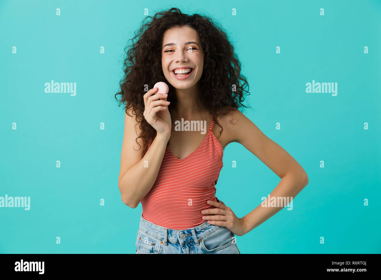 Photo de charmante femme 20s avec les cheveux bouclés de sourire et de manger biscuit macaron isolé sur fond bleu Banque D'Images