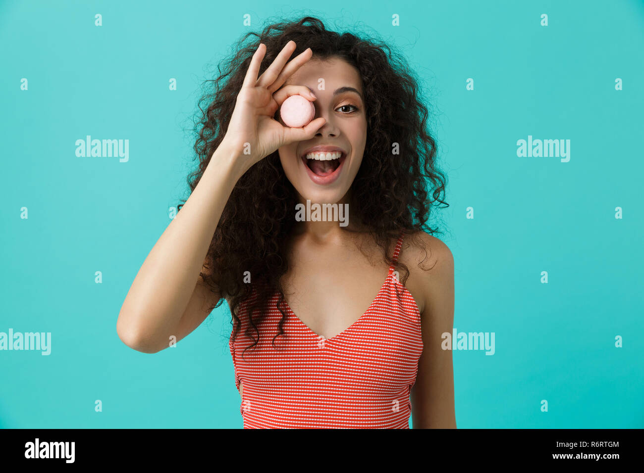 Photo de portrait femme 20s avec les cheveux bouclés de sourire et de manger biscuit macaron isolé sur fond bleu Banque D'Images