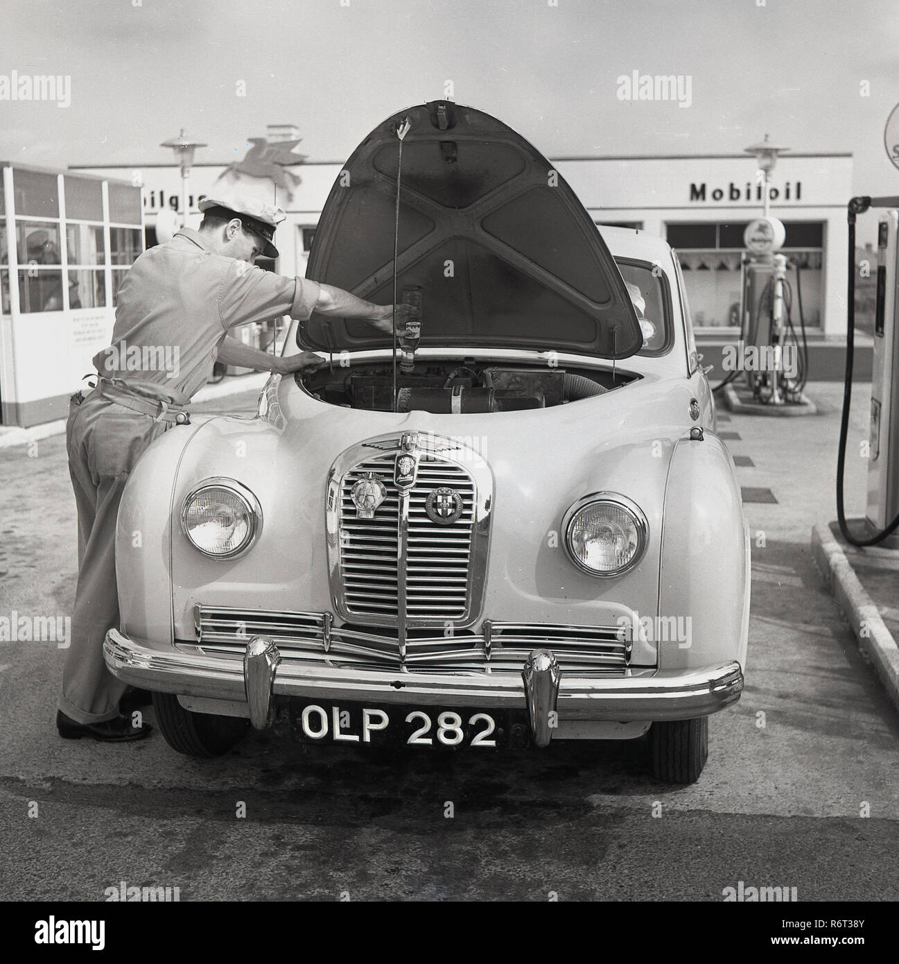 Années 1950, historiques, une automobile de l'époque à la pompe à carburant, avec son capot ouvert étant donné un service d'huile par un préposé garage en uniforme, England, UK. En cette ère d'or de l'automobile, de nombreux garages ou ce qu'on appelait le service des stations spécialisées a bord qui ainsi que le ravitaillement de la voiture vous offrent également de l'entretien de routine et vérifier les véhicules les hydrocarbures, l'eau et pression des pneus. Banque D'Images