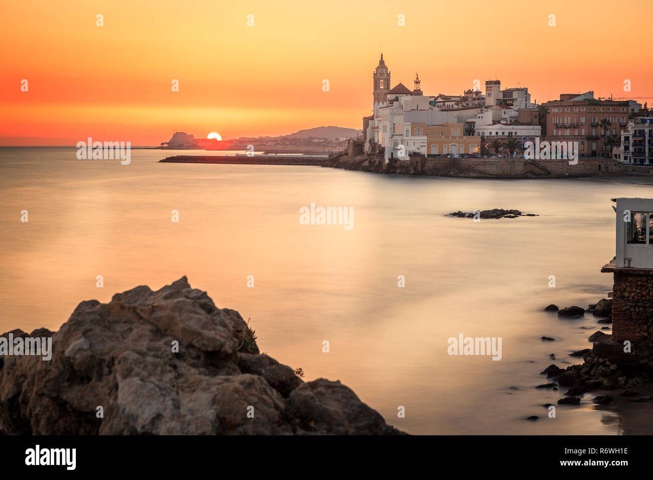 C'est un coucher de soleil avec des pierres au premier plan, la mer Méditerranée par blurried une faible vitesse d'obturation et la ville de Sitges en arrière-plan. Banque D'Images