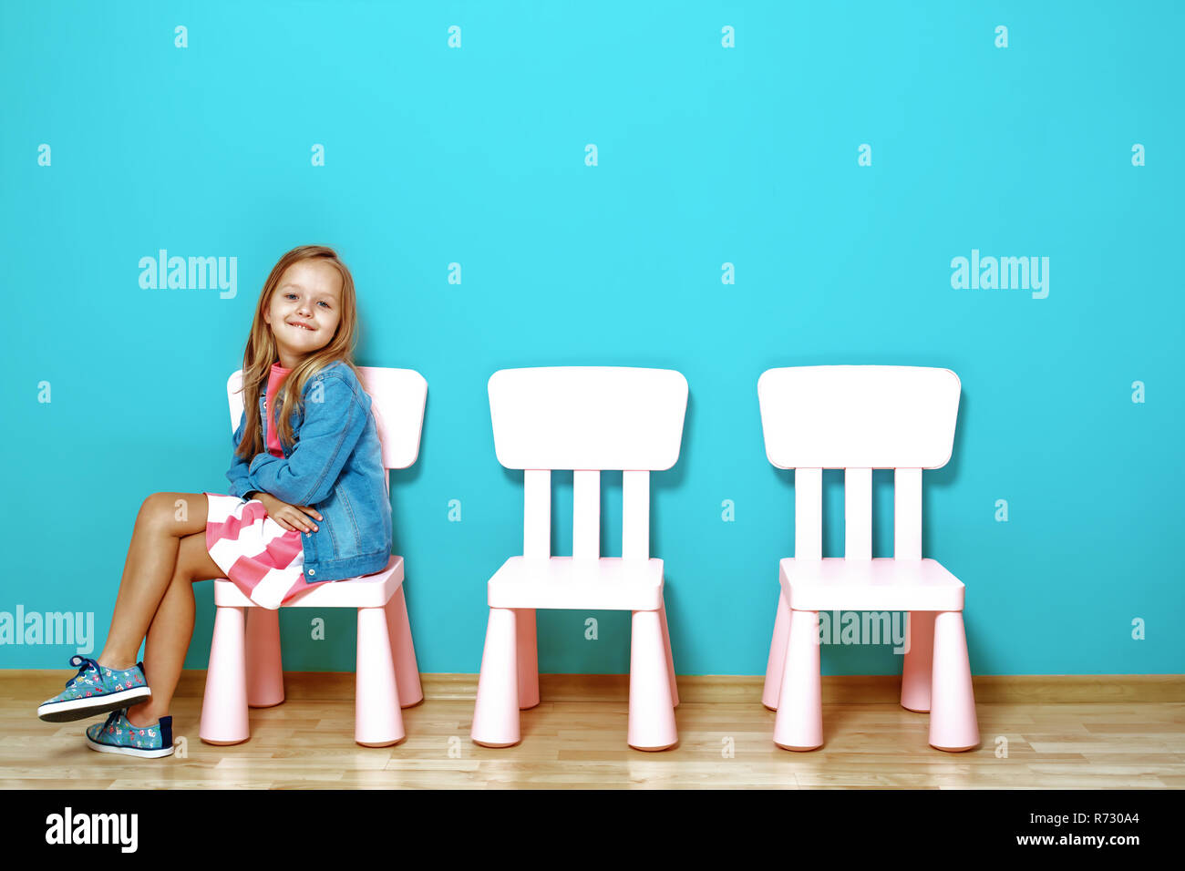 Heureux petit enfant fille est assis sur la chaise dans le contexte de la mur bleu. A proximité se trouvent des chaises vides. La notion de bonheur, de l'enfance Banque D'Images