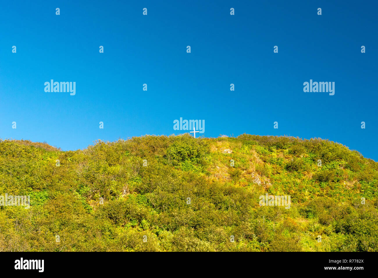 Petite croix chrétienne sur un sommet de montagne verte contre un vaste ciel bleu clair. Banque D'Images