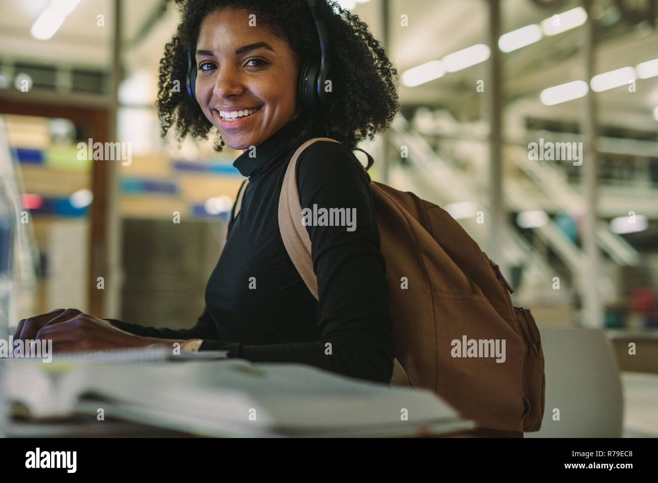 Happy african american woman student with laptop and books sitting at table smiling at camera. Female student studying in library. Banque D'Images