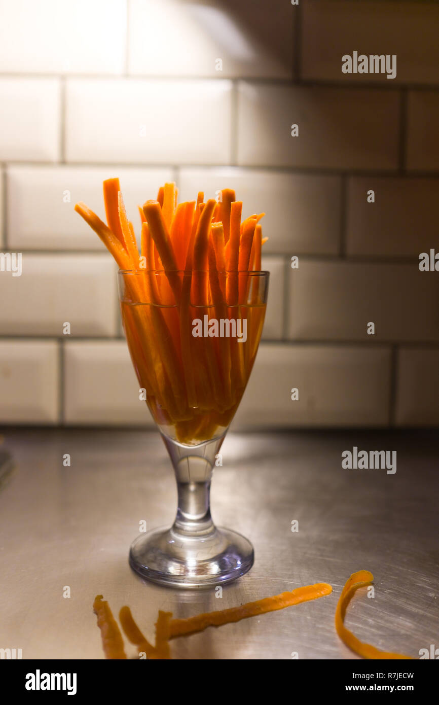 Bâtonnets de carotte crue dans un verre a pied d'eau à côté de Peel sur une  surface de travail en acier inoxydable dans une cuisine domestique avec  carreaux de métro en arrière-plan