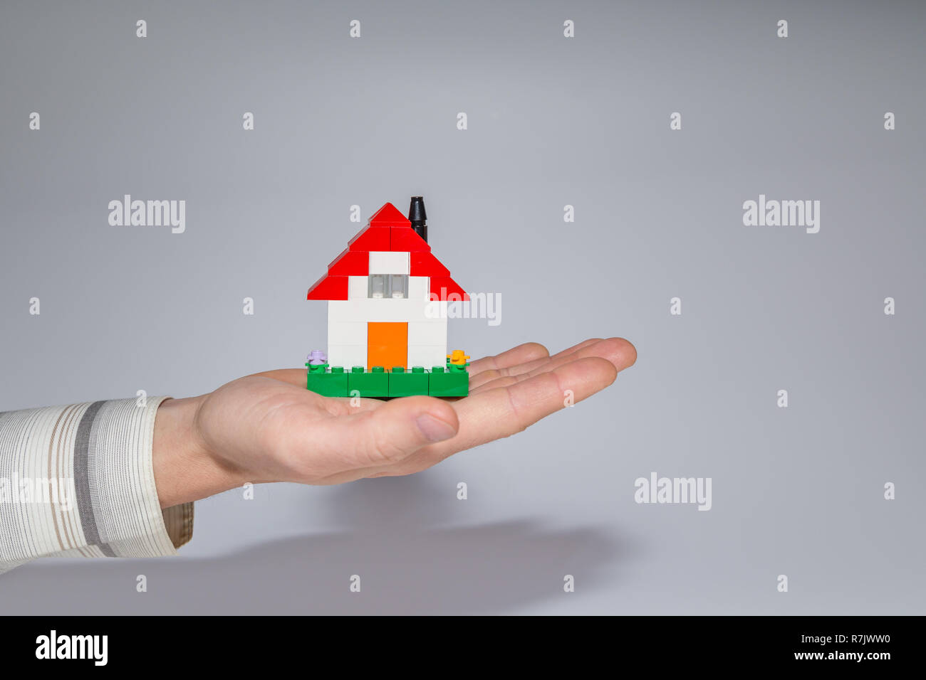 Homme tenant une petite maison faite de briques de construction de jouets sur sa paume. Fond gris. Photo de concept immobilier maison ou propriété. Banque D'Images