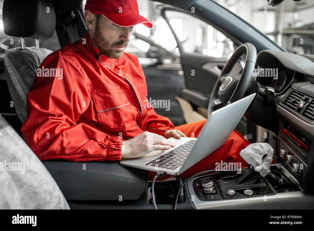 Mécanicien Auto en uniforme rouge diagnostic voiture avec ordinateur assis sur le siège conducteur de la voiture publique Banque D'Images