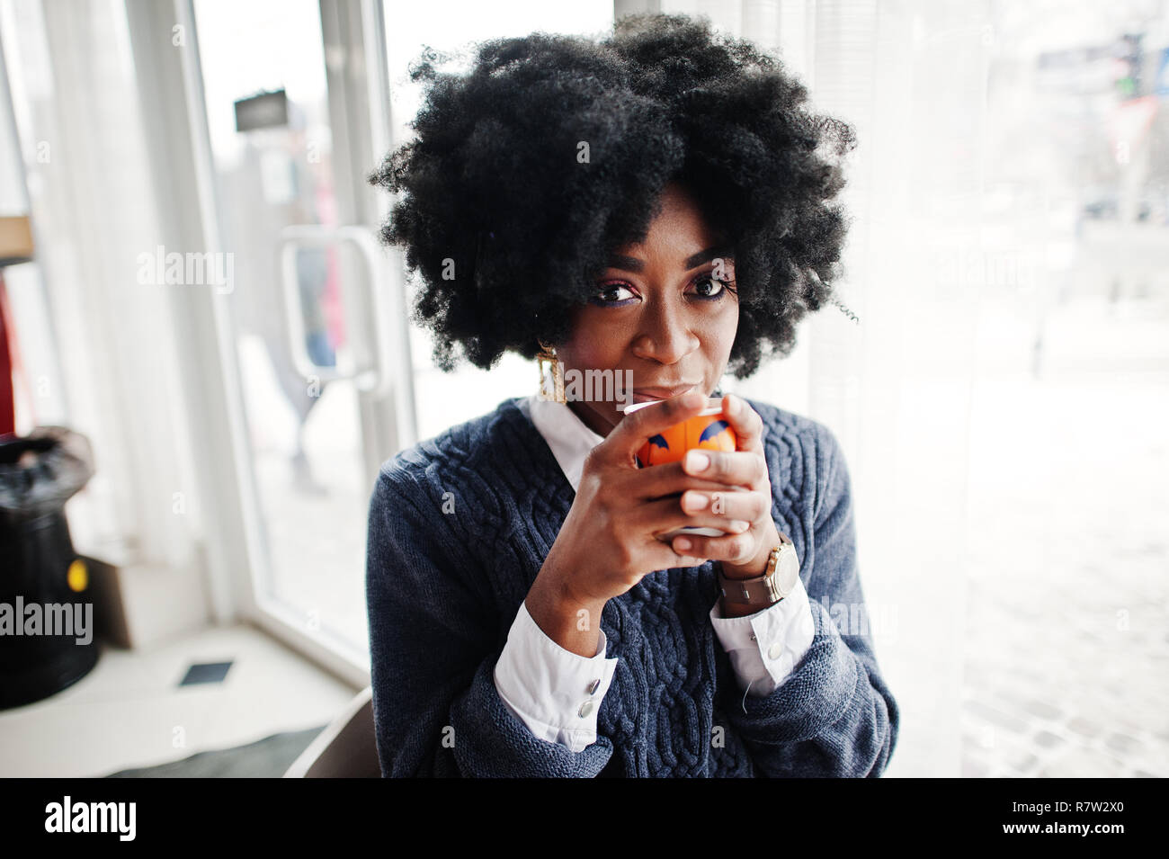 Cheveux bouclés african american woman usure sur sweater posés au café avec une tasse de thé ou de café. Banque D'Images