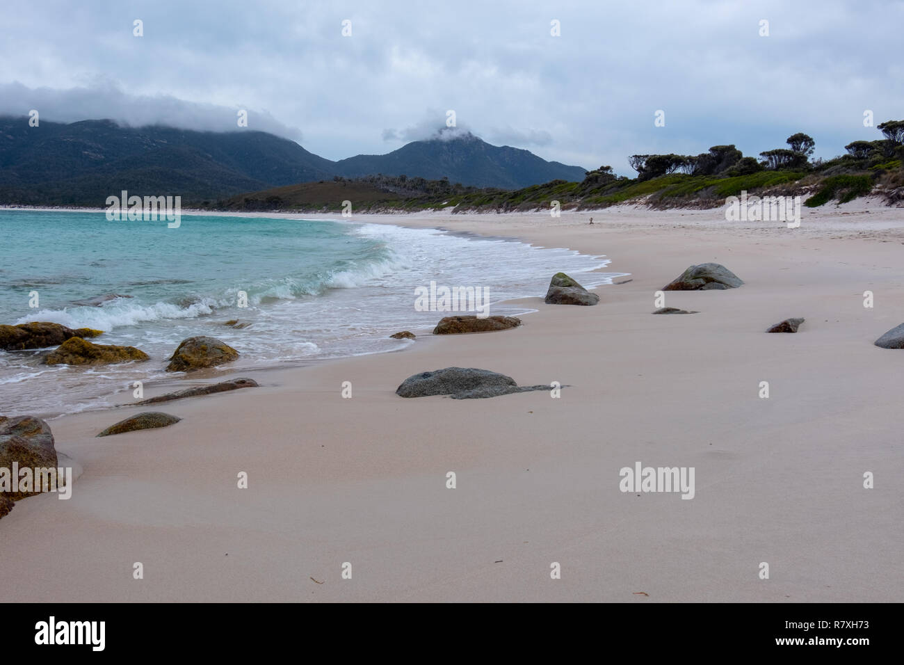Plage de Wineglass Bay, parc national de Freycinet, la Tasmanie avec des pierres à l'avant-plan et d'une mer bleue sur un jour nuageux Banque D'Images