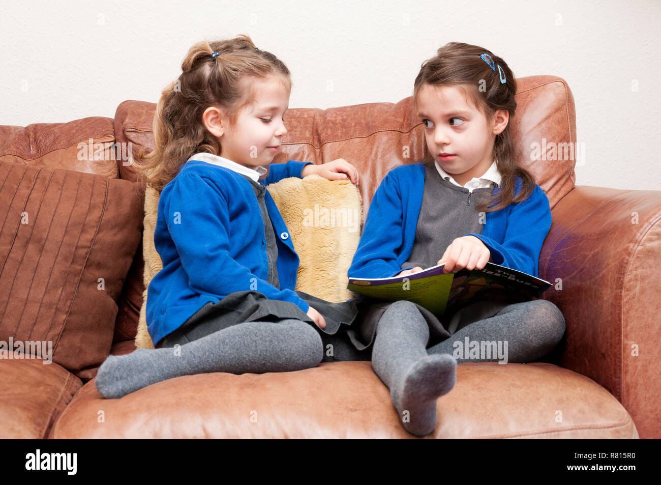 Les jeunes sœurs en uniforme de l'école primaire la lecture d'un livre ensemble, assis sur un canapé, Royaume-Uni Banque D'Images