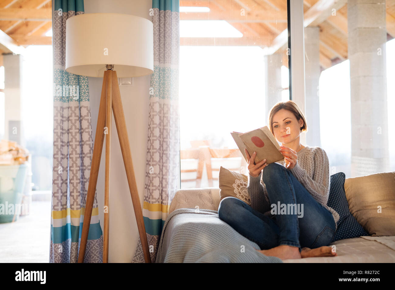 Une jeune femme assise à l'intérieur sur un canapé à la maison, la lecture d'un livre. Banque D'Images