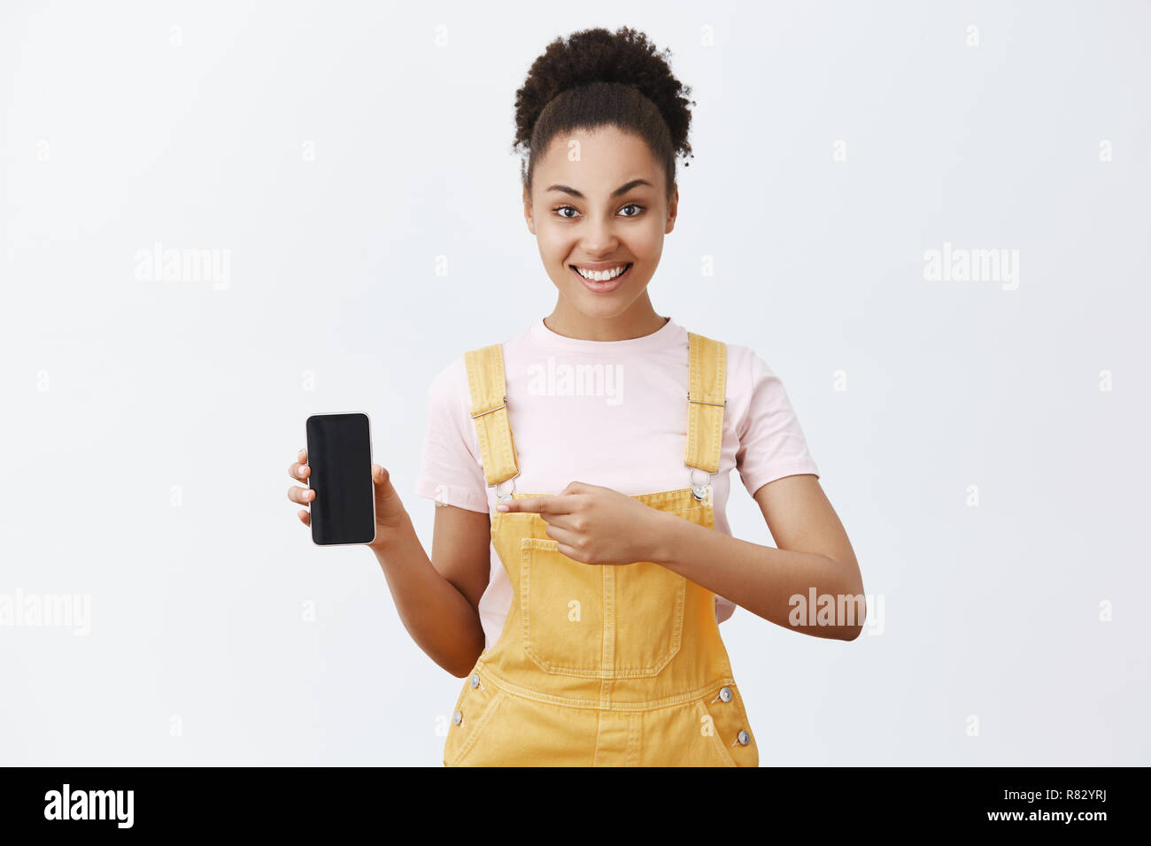 Cliquez sur ce bouton. Carefree friendly-à la femme africaine en étudiant la mode salopette jaune, holding smartphone et pointage du doigt d'index à l'appareil, tout sourire, suggérant great app Banque D'Images