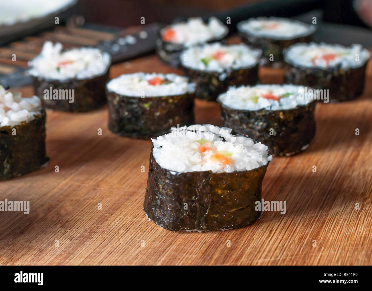 Chumaki rouleaux de sushi au thon, saumon, riz, concombre, d'avocat et d'algue Nori sur une planche en bois avec des ustensiles en arrière-plan flou. Banque D'Images