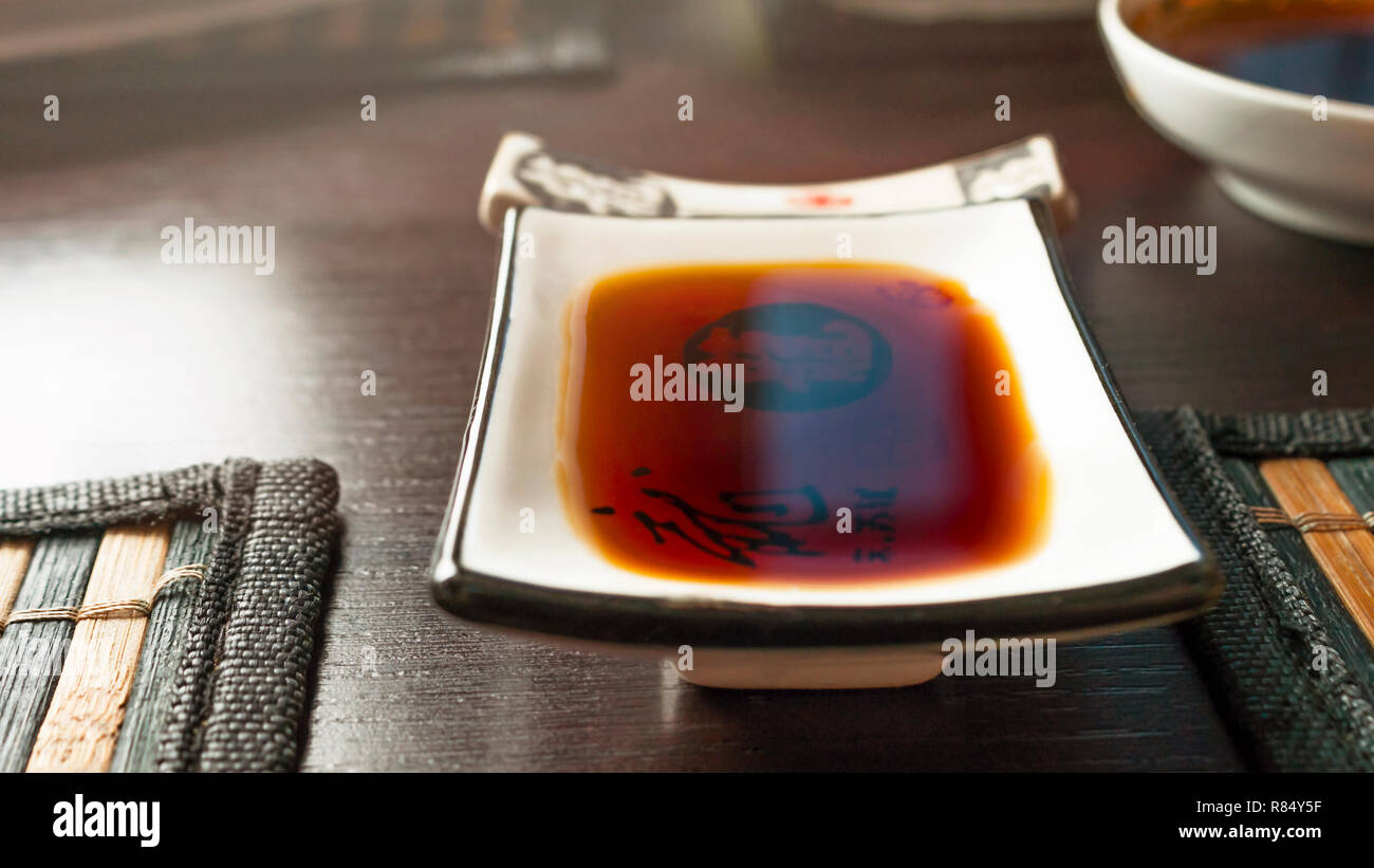Sauce soja plat rectangulaire inclinée sur une table de salle à manger en bambou foncé avec des tapis. Chopstick reste, Cuve en arrière-plan flou. Lueur chaude. Banque D'Images