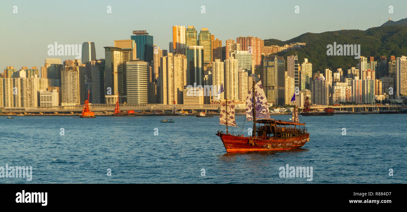 Jonque Chinoise avec Mauve et blanc vogue sur l'eau dans le port de Hong Kong au coucher du soleil avec des bâtiments en arrière-plan. Banque D'Images