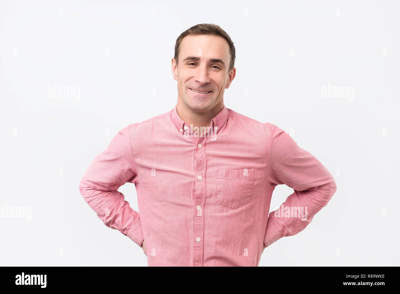 Bel homme italien portant chemise rose avec léger sourire posant contre fond blanc Banque D'Images