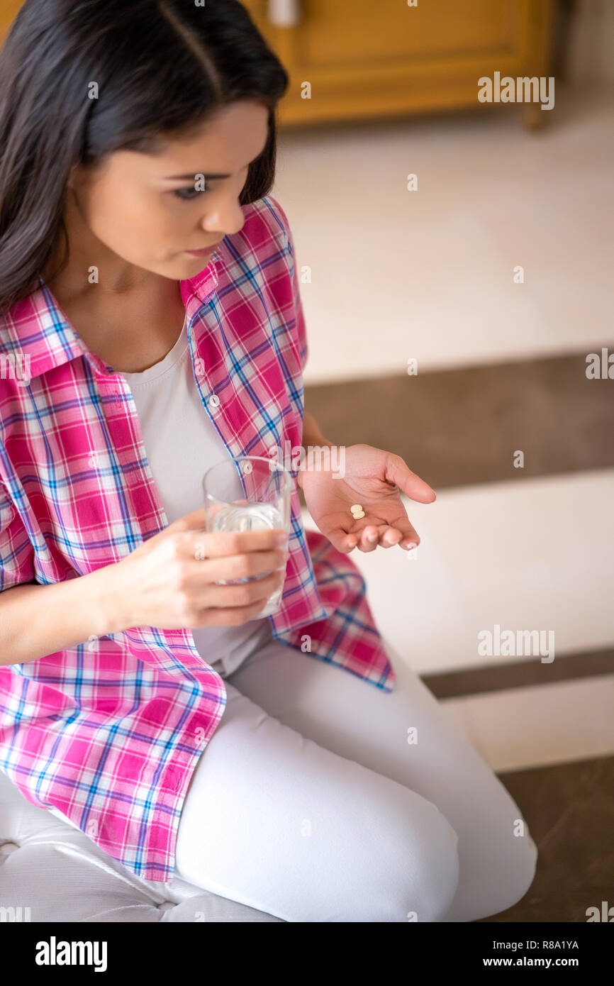 Contrarié Femme assise sur le canapé et détient les comprimés et un verre d'eau. La médecine. Le traitement. Banque D'Images