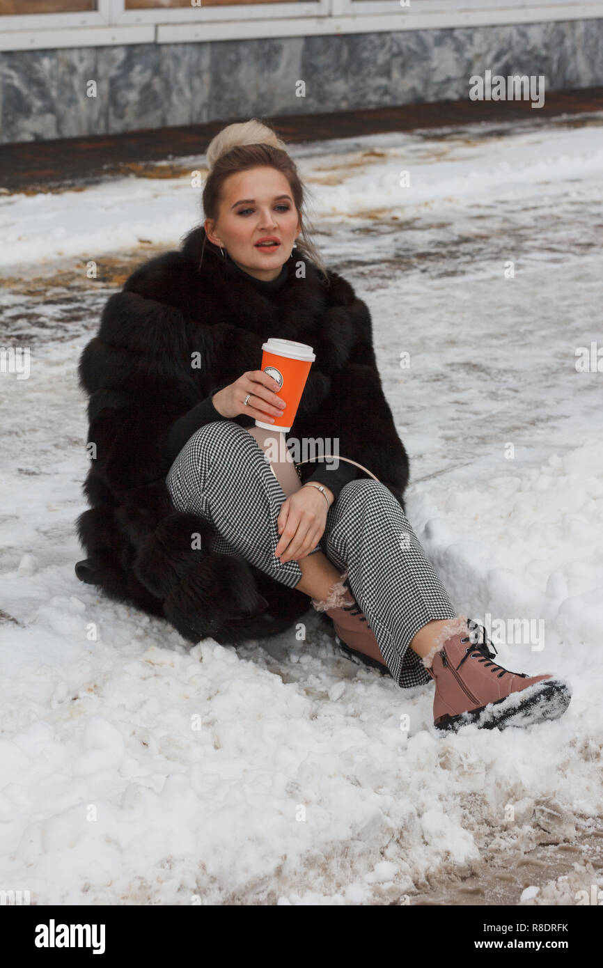 Girl sitting on a de la neige dans la ville le trottoir couvert de neige. L'humeur d'hiver. Vie, tasse de café, le vison vêtements de fourrure. Banque D'Images