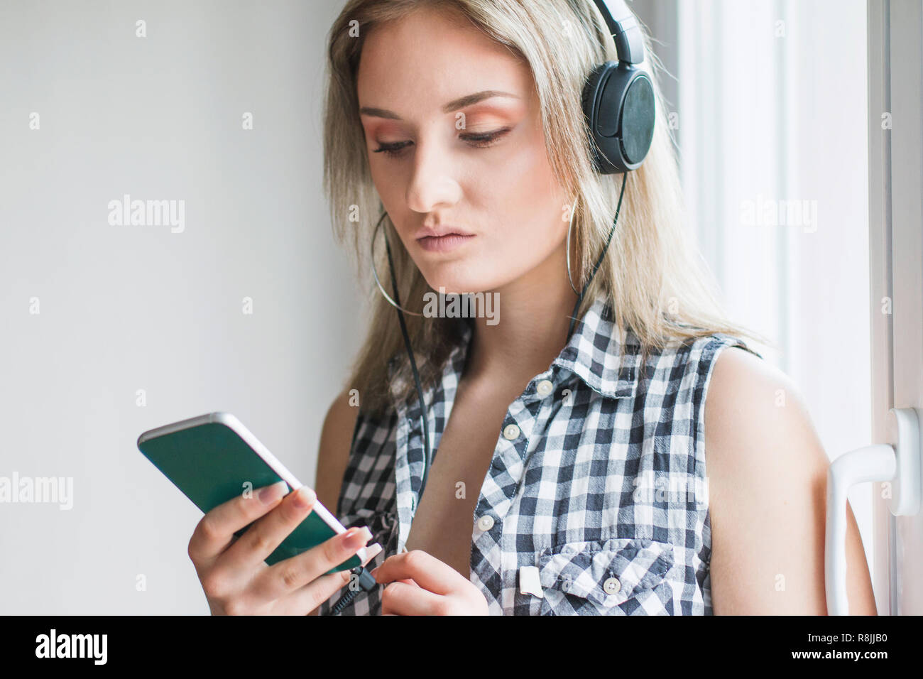 Belle jeune fille moderne cheveux longs en chemise à carreaux avec des écouteurs et smart phone assis à la maison et la musique d'origami, blanc Banque D'Images