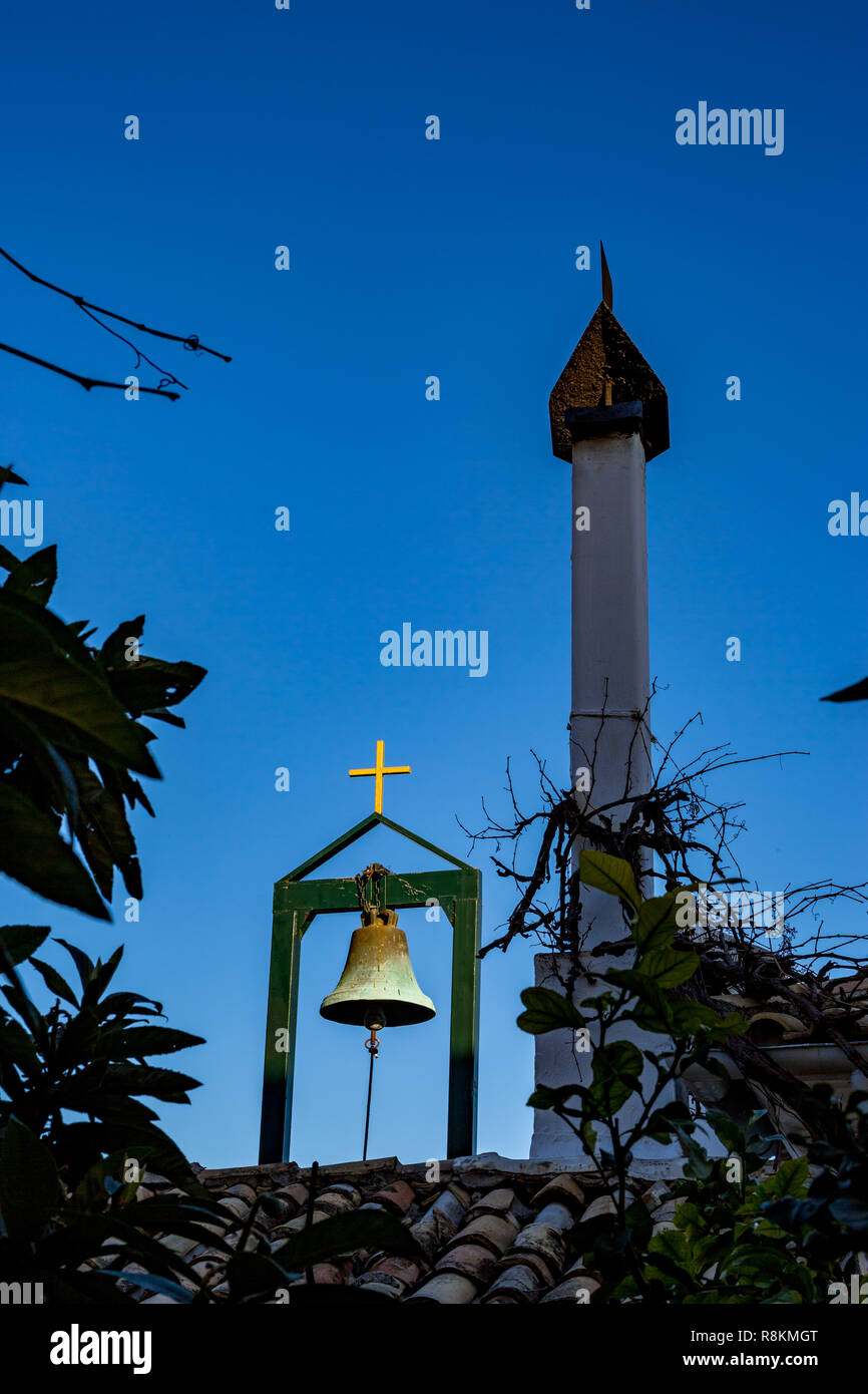 Monastère grec-orthodoxe détails toit, cloche en laiton sur socle, croix d'or et haute cheminée au toit en tuiles rouges dans le ciel bleu clair jour de printemps Banque D'Images