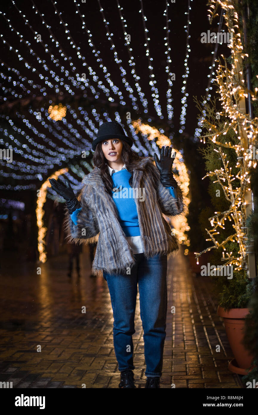 Jolie fille aux cheveux noirs portant un manteau de fourrure, jeans, haut bleu et un chapeau noir, sourire, posant avec des flocons de lumières de Noël à l'extérieur nuit ti Banque D'Images