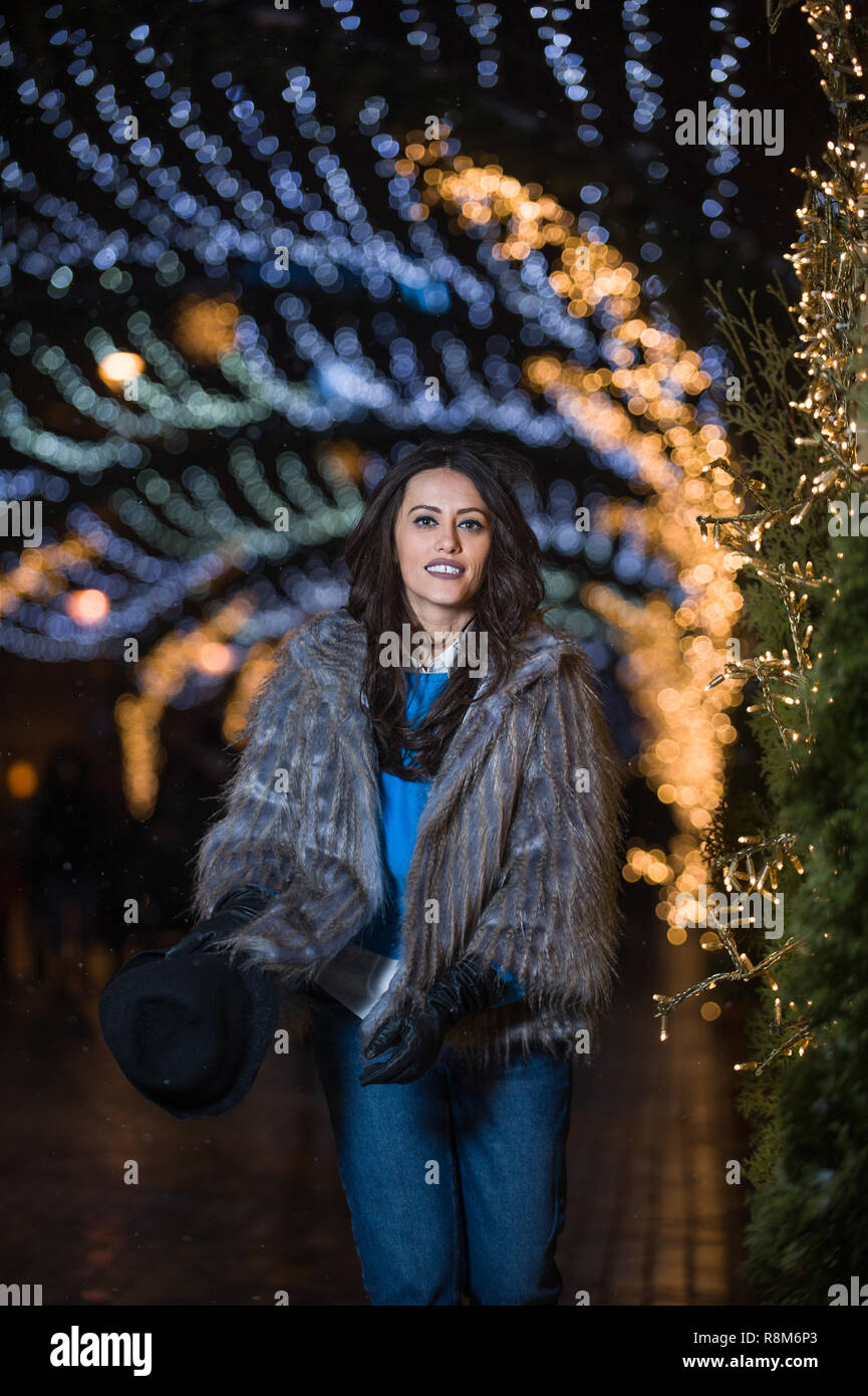 Jolie fille aux cheveux noirs portant un manteau de fourrure, jeans, haut bleu et un chapeau noir, sourire, posant avec des flocons de lumières de Noël à l'extérieur nuit ti Banque D'Images