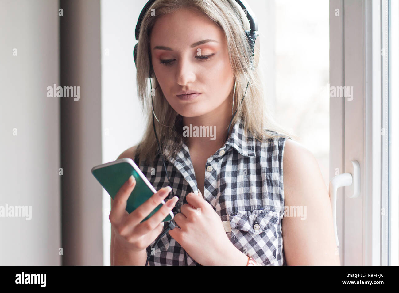 Belle jeune fille moderne cheveux longs en chemise à carreaux avec des écouteurs et smart phone assis à la maison et la musique d'origami, blanc Banque D'Images