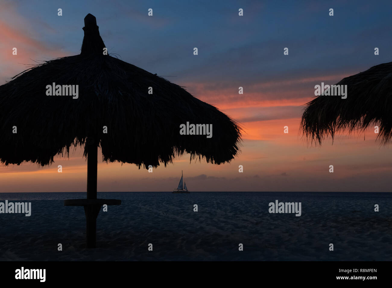 Eagle Beach Aruba au coucher du soleil - un voilier apparaît sous des parasols en chaume avec rose et bleu ciel sur la rive nord-ouest d'Aruba Banque D'Images