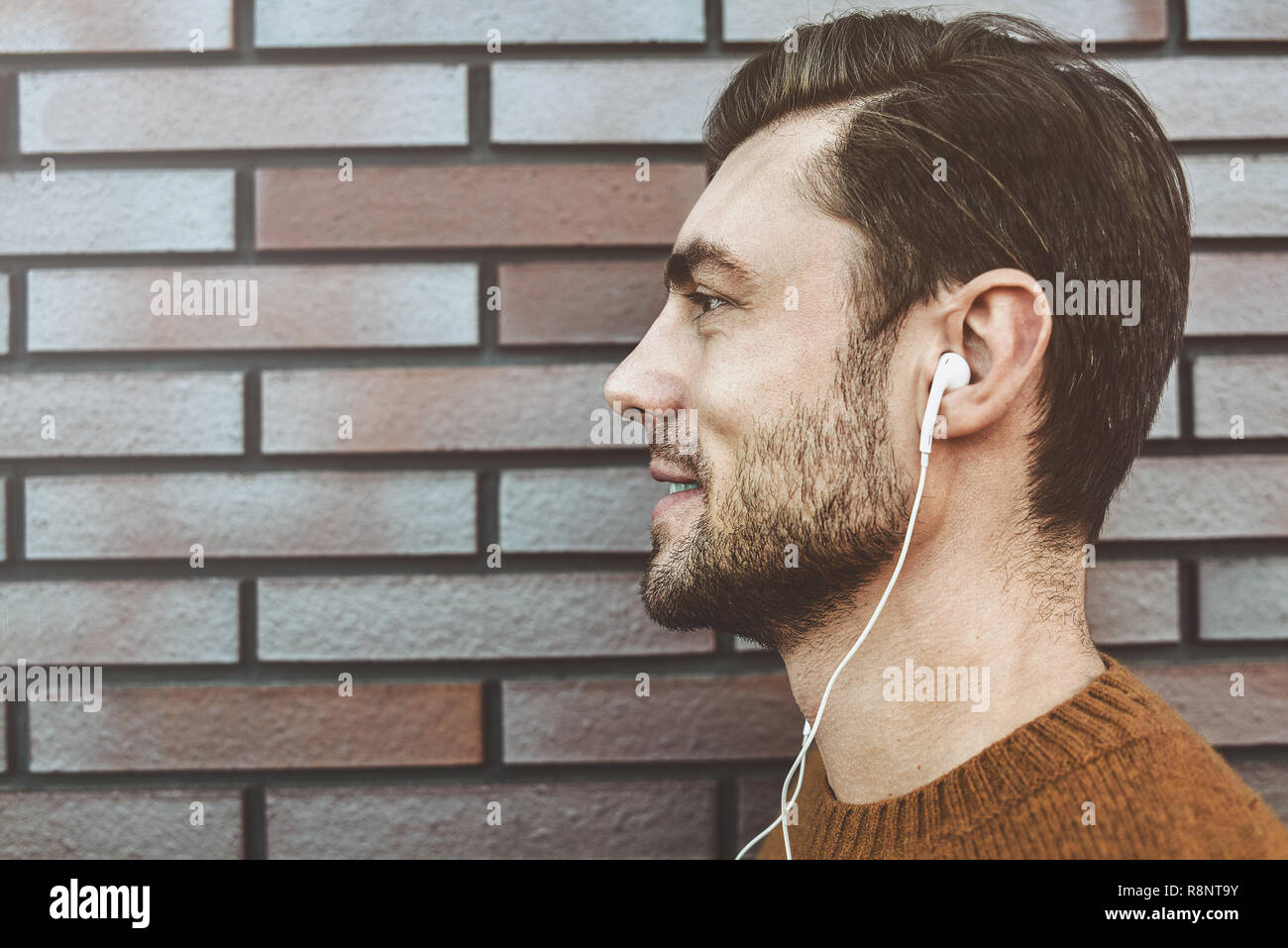 Photo de beau jeune homme élégant avec des pinceaux en soie de l'article à l'extérieur. Homme portant un chandail brun. Smiling man listening to music on headphones et leanin Banque D'Images