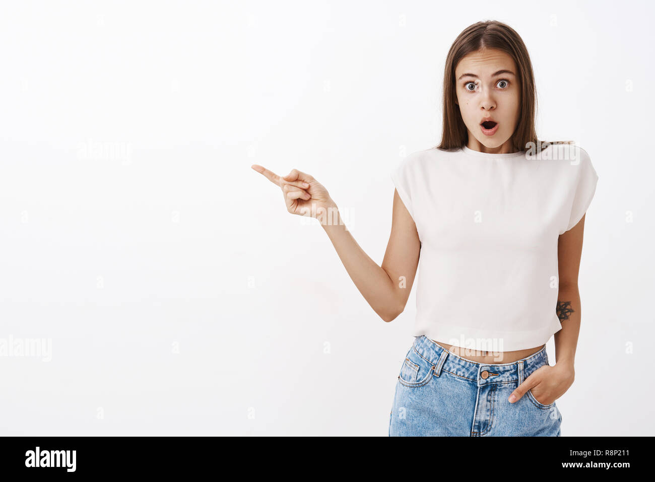 Remise en question et choqué girl pointant vers la gauche de l'ouverture de la bouche de surprise et de stupéfaction holding hand in pocket et permanent à partir de scène incroyable caméra secoua sur fond gris Banque D'Images