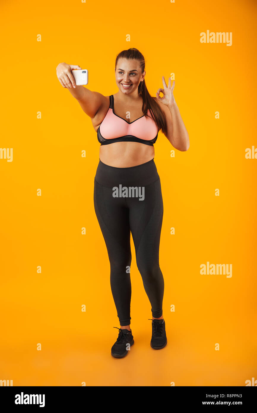 Full Length portrait of smiling woman in chubby soutien-gorge sportif prendre photo selfies sur téléphone mobile sur fond jaune isolé Banque D'Images