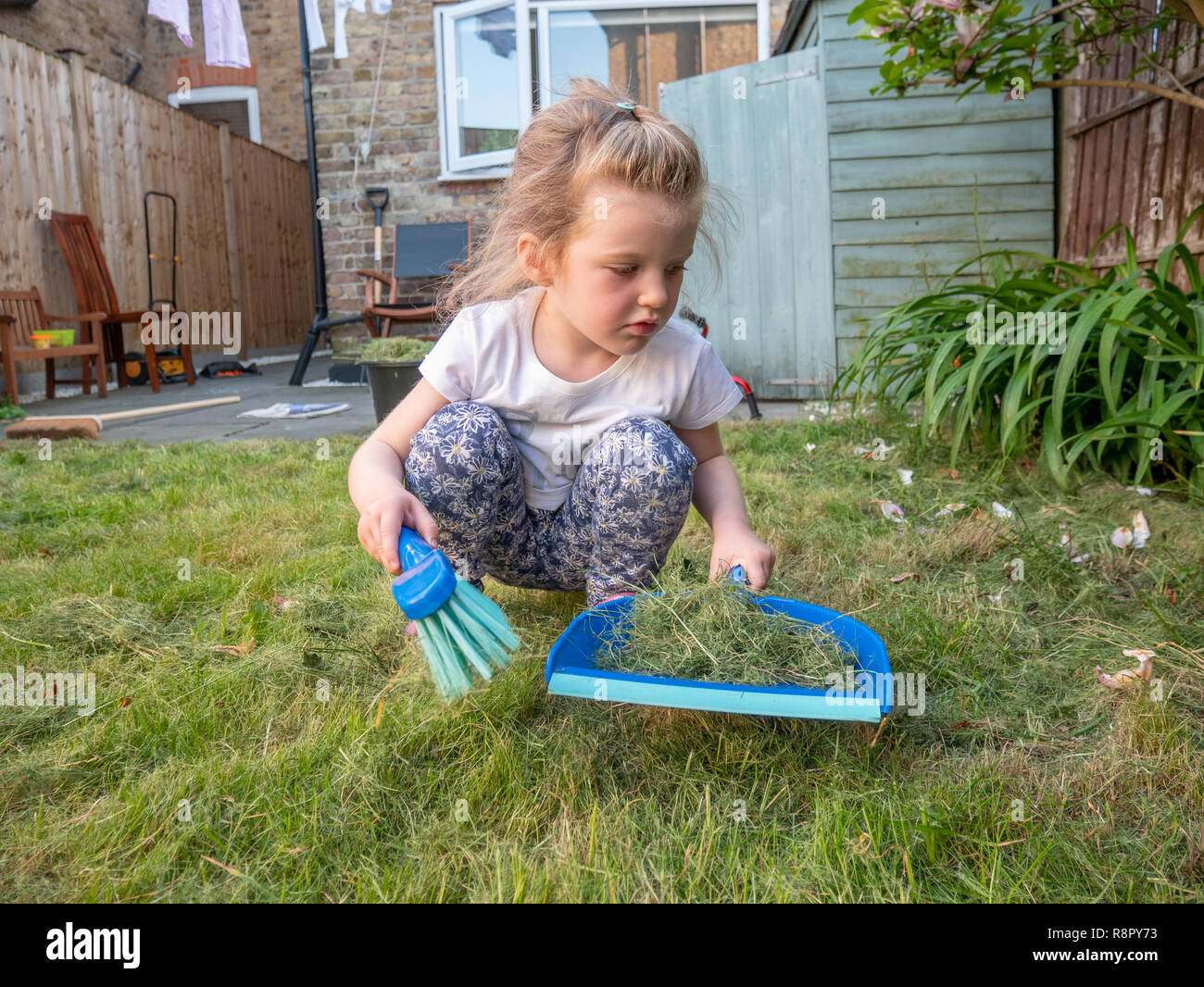 Trois ans, fille, balayant le haut de l'herbe coupée dans le jardin arrière, London, UK Banque D'Images