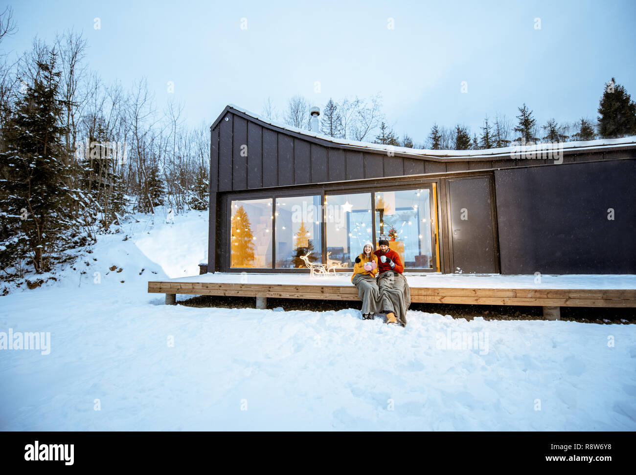 Jeune couple de préchauffage avec plaid et breuvages assis sur la terrasse de la maison moderne dans les montagnes durnig les vacances d'hiver. Large vue landsacpe Banque D'Images