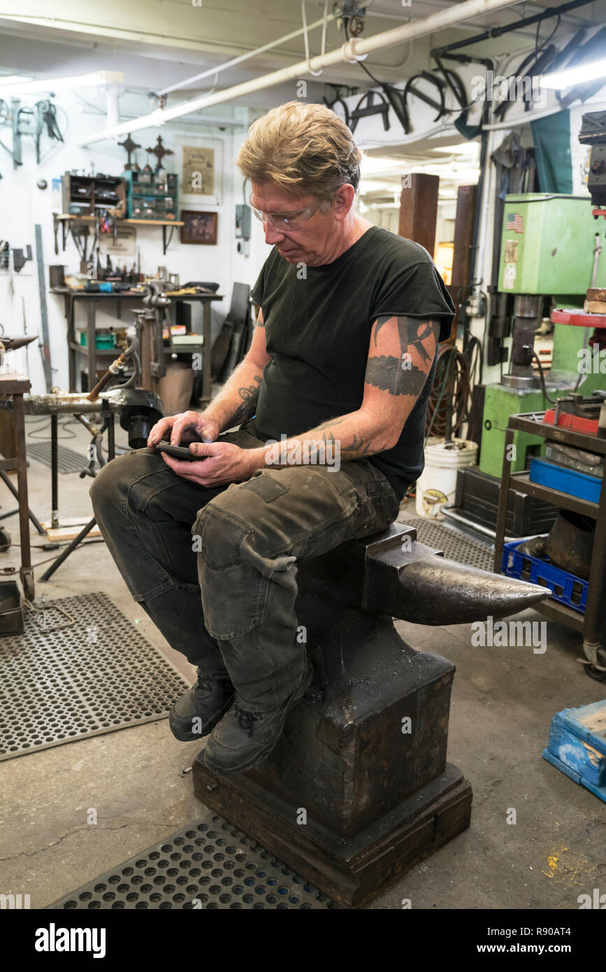 Contrôle de forgeron mâle son téléphone pour les messages tout en étant assis sur une enclume dans son studio. Banque D'Images