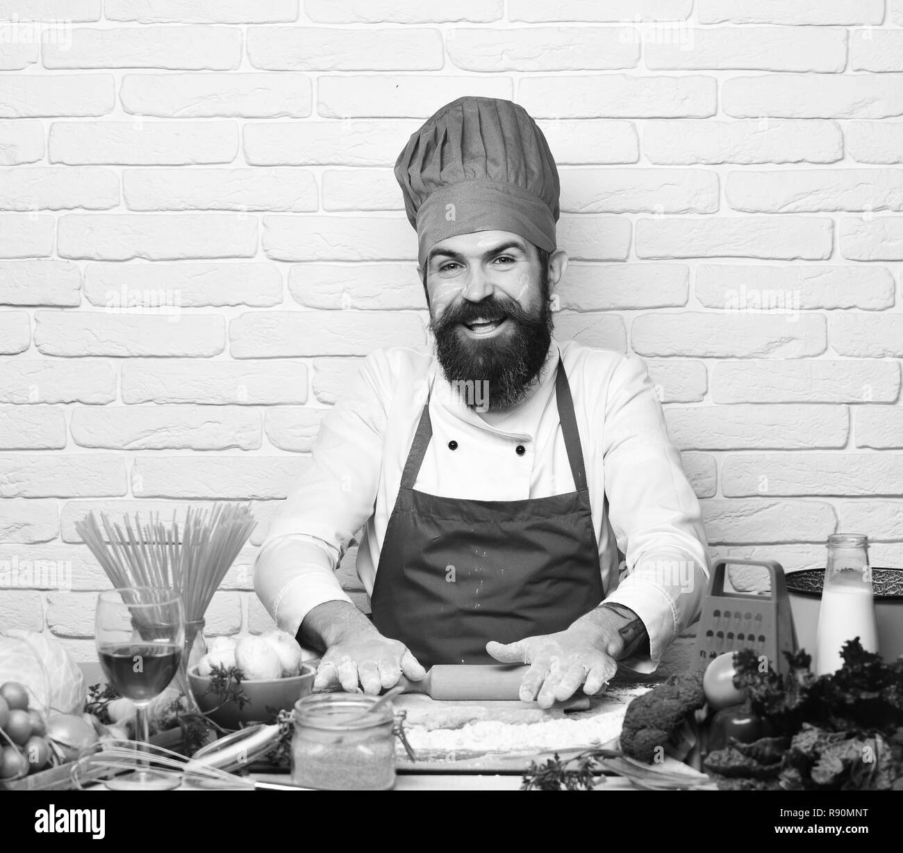 Le chef fait la pâte. Concept de la cuisine italienne. Homme avec barbe sort en pâte sur mur de brique blanc arrière-plan. Cook avec visage rieur en bourgogne est uniforme par table avec des légumes et des ustensiles de cuisine. Banque D'Images