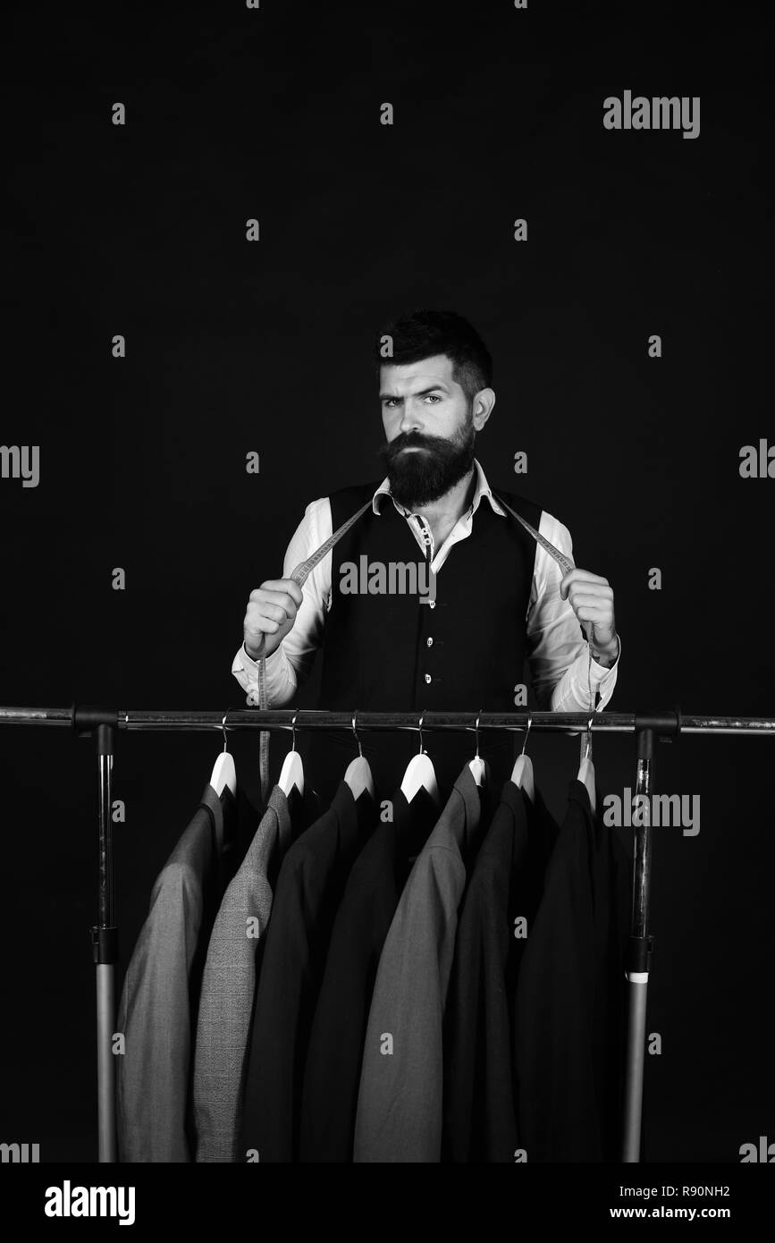 Avec l'homme à barbe sont acquis par porte manteau. Designer près de cintres. La mode et le style individuel concept. Adapter avec visage sérieux est titulaire d'un ruban à mesurer près de vestes sur mesure sur fond noir Banque D'Images