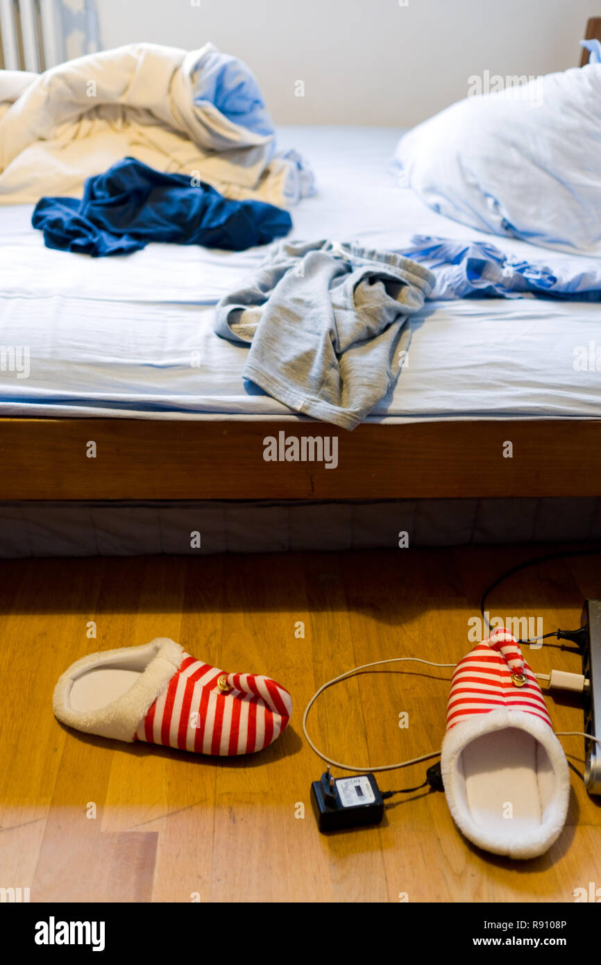 Vue de deux elf de chaussons, d'un chargeur de téléphone portable et d'une rallonge sur plancher en bois par un lit défait avec les vêtements dans une chambre à coucher des adolescents de sexe masculin Banque D'Images