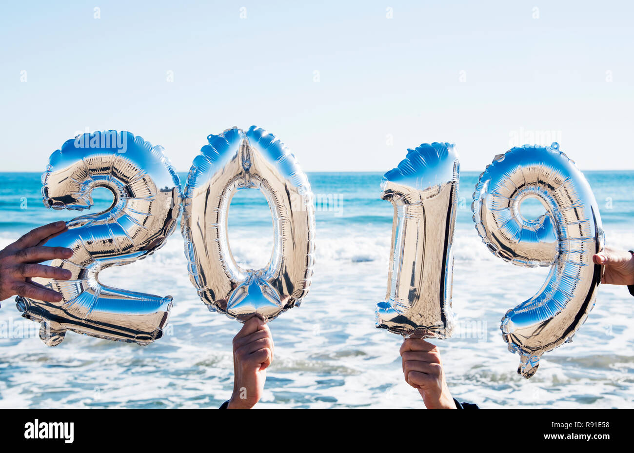 Les mains des gens à l'extérieur tenant quelques ballons en forme de numéro argenté formant le nombre 2019, comme la nouvelle année, et de l'océan en arrière-plan Banque D'Images