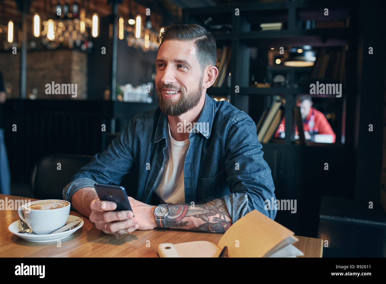 Jeune homme barbu, vêtu d'une chemise en jean, assis à table dans un café et d'utiliser votre smartphone. L'homme à l'aide de gadget. Banque D'Images