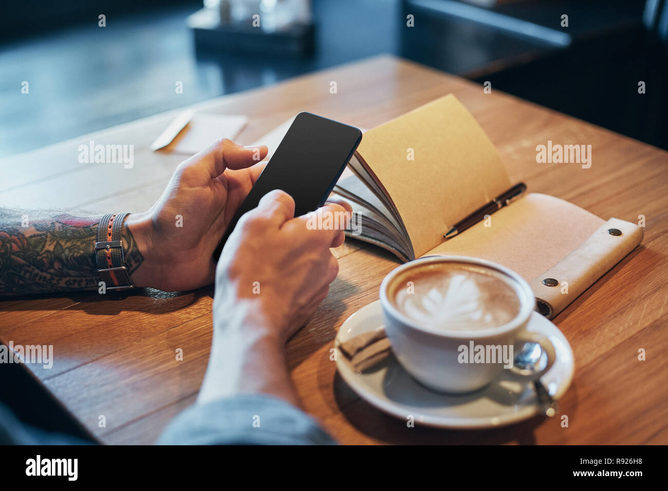 Les mains de l'homme en chemise en jean glisser avec le doigt sur l'écran de son téléphone intelligent, près de tasse à café sur table en bois Banque D'Images