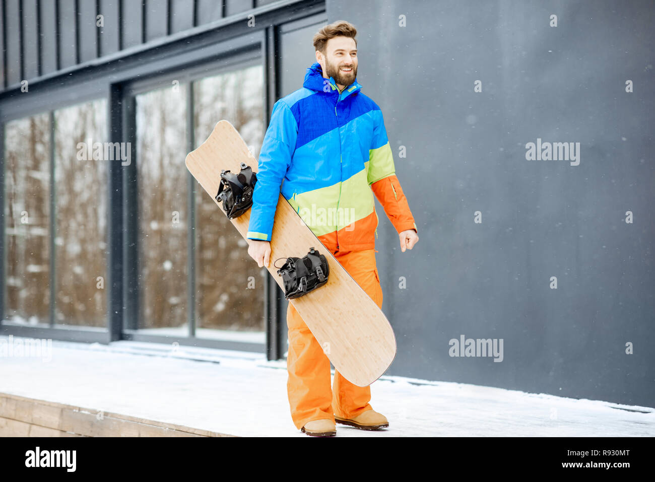 Homme En Combinaison De Ski Avec Snowboard Près De La Maison Ou De L'hôtel  Moderne Dans Les Montagnes