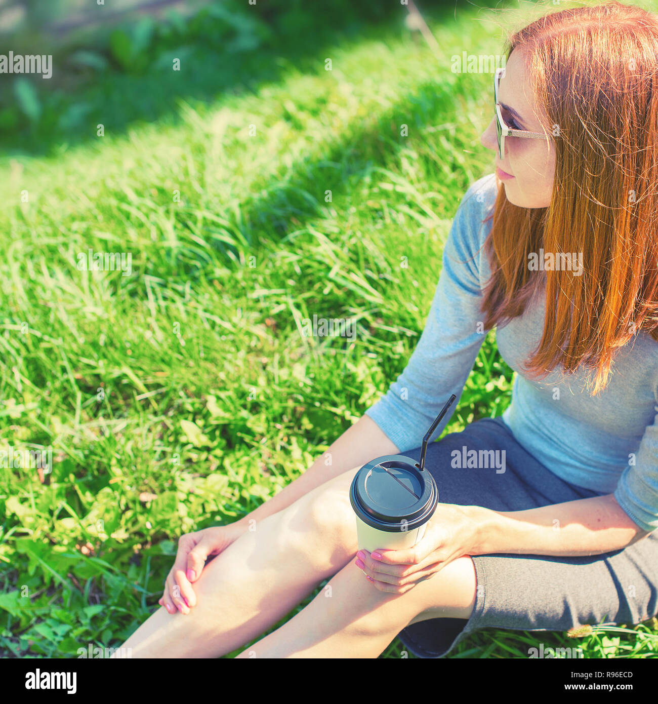 Belle fille aux cheveux roux assis sur l'herbe et boire du café dans une tasse de papier. Petit-déjeuner de style de vie dans le parc Banque D'Images