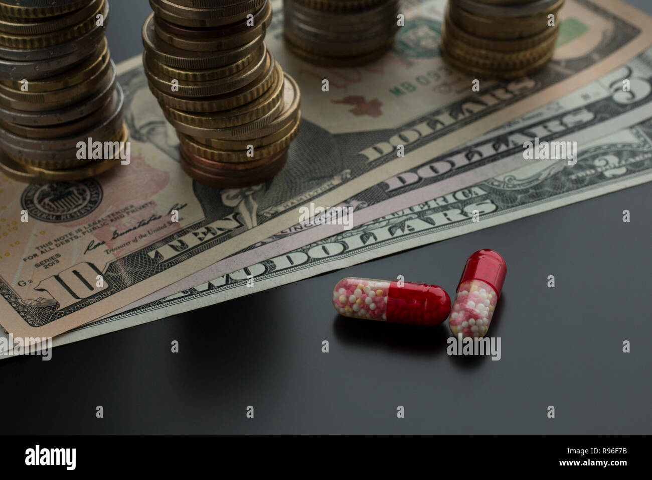 Deux comprimés ou capsules rouges contre des dollars et des piles de pièces de monnaie sur l'arrière-plan. Concept de traitement coûteux Banque D'Images