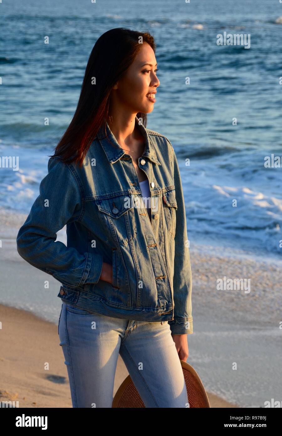 Une jeune femme, portant une veste en denim et jeans, est installé dans un cadre paisible en fin d'après-midi promenade sur la plage. Banque D'Images