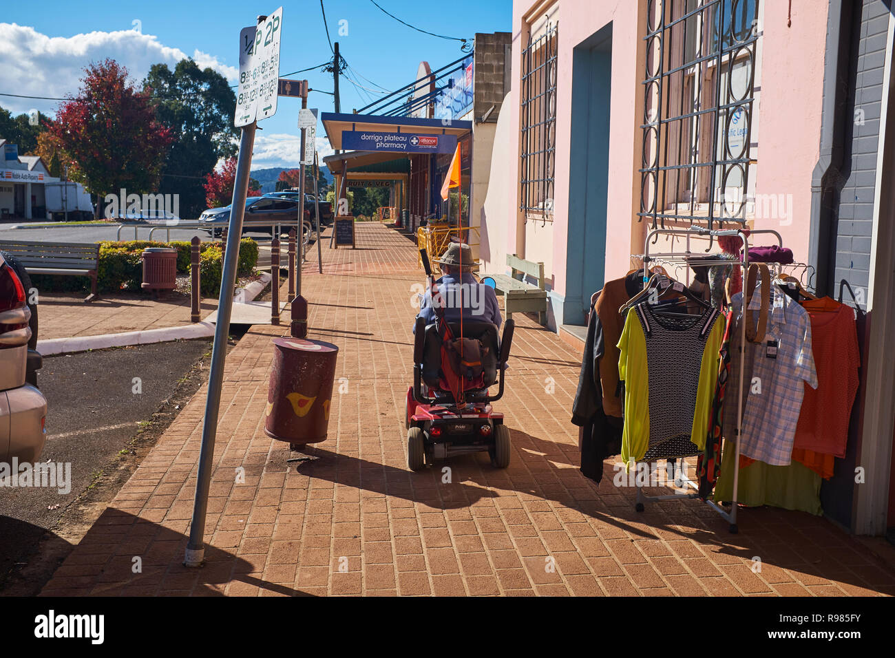 La vue arrière d'une personne qui conduit un scooter de mobilité sur le trottoir passant un magasin qui a des vêtements pour la vente à l'extérieur, Dorrigo, NSW, Australie Banque D'Images