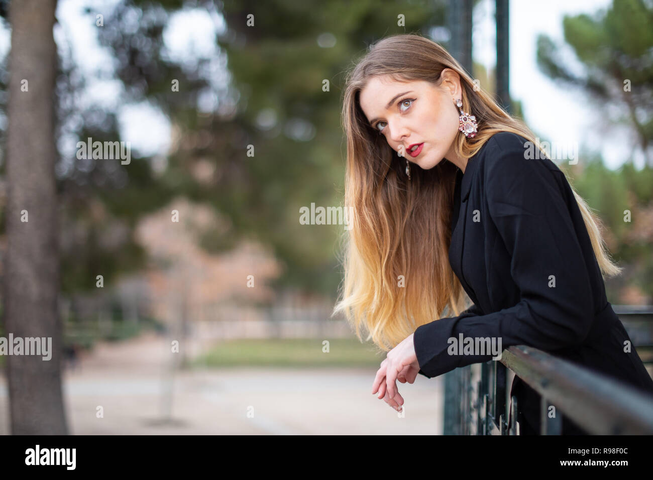 Vue latérale du séduisant jeune femme en costume élégant leaning on fence et à l'écart en se tenant sur le fond flou de park Banque D'Images