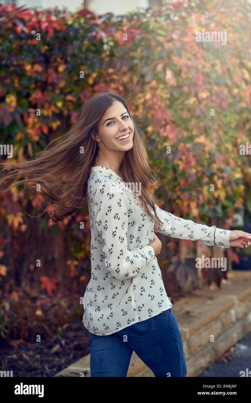 Carefree woman rejetant ses cheveux alors qu'elle regarde en arrière de l'appareil photo avec un sourire plein de vivacité en face de feuillage automne coloré Banque D'Images