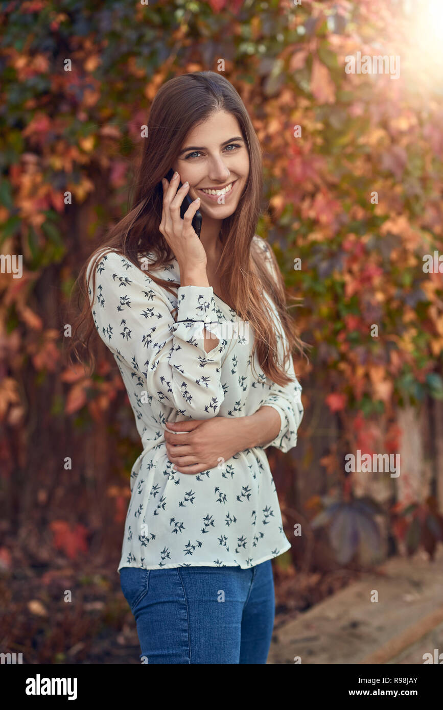 Souriante jeune femme chat sur un mobile à l'automne, debout devant un réducteur coloré souriant joyeusement à l'appareil photo avec sun flare Banque D'Images