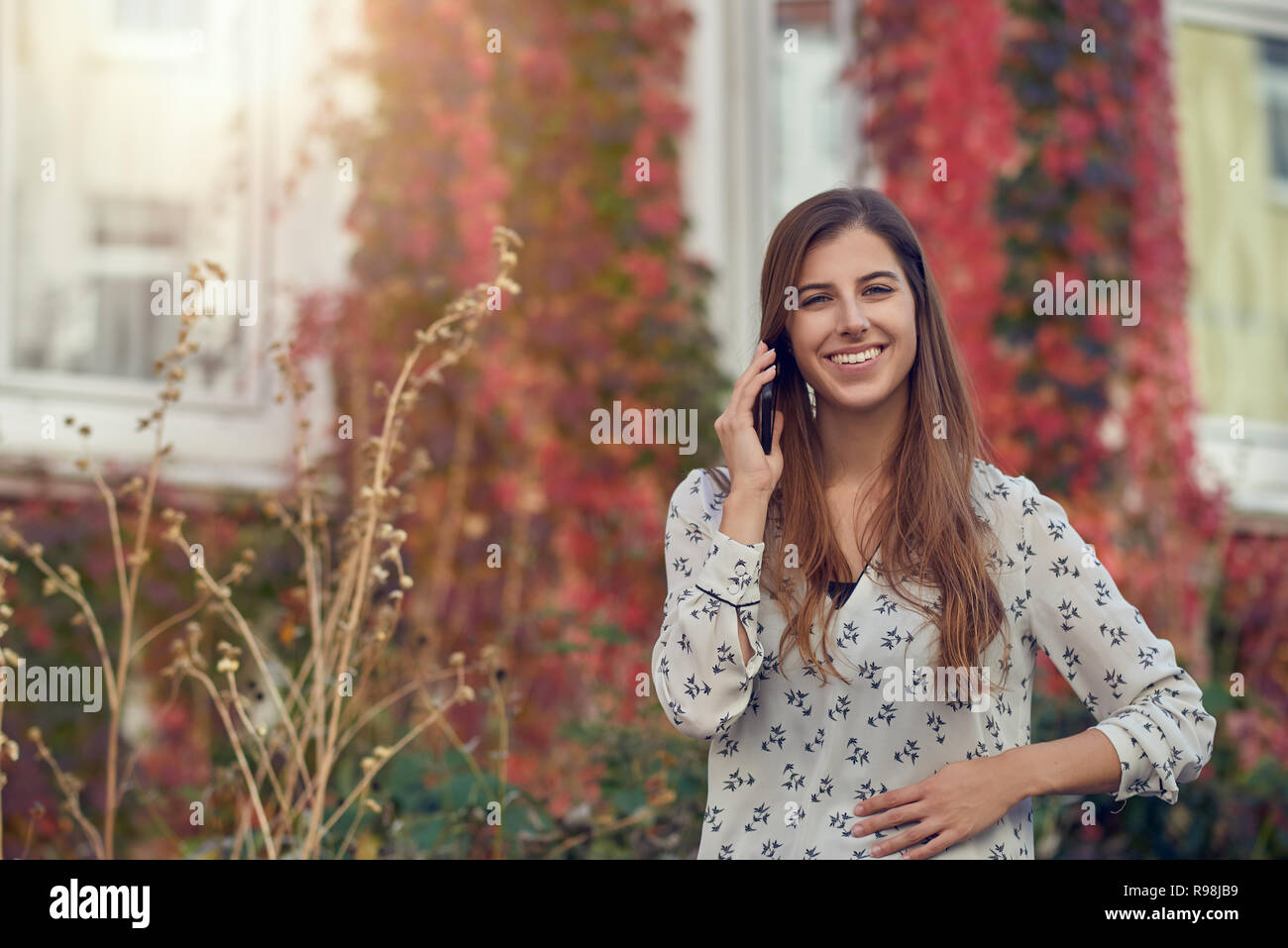 Souriante jeune femme chat sur un mobile à l'automne debout devant une maison blanche avec réducteur coloré souriant joyeusement à l'appareil photo avec fla Banque D'Images